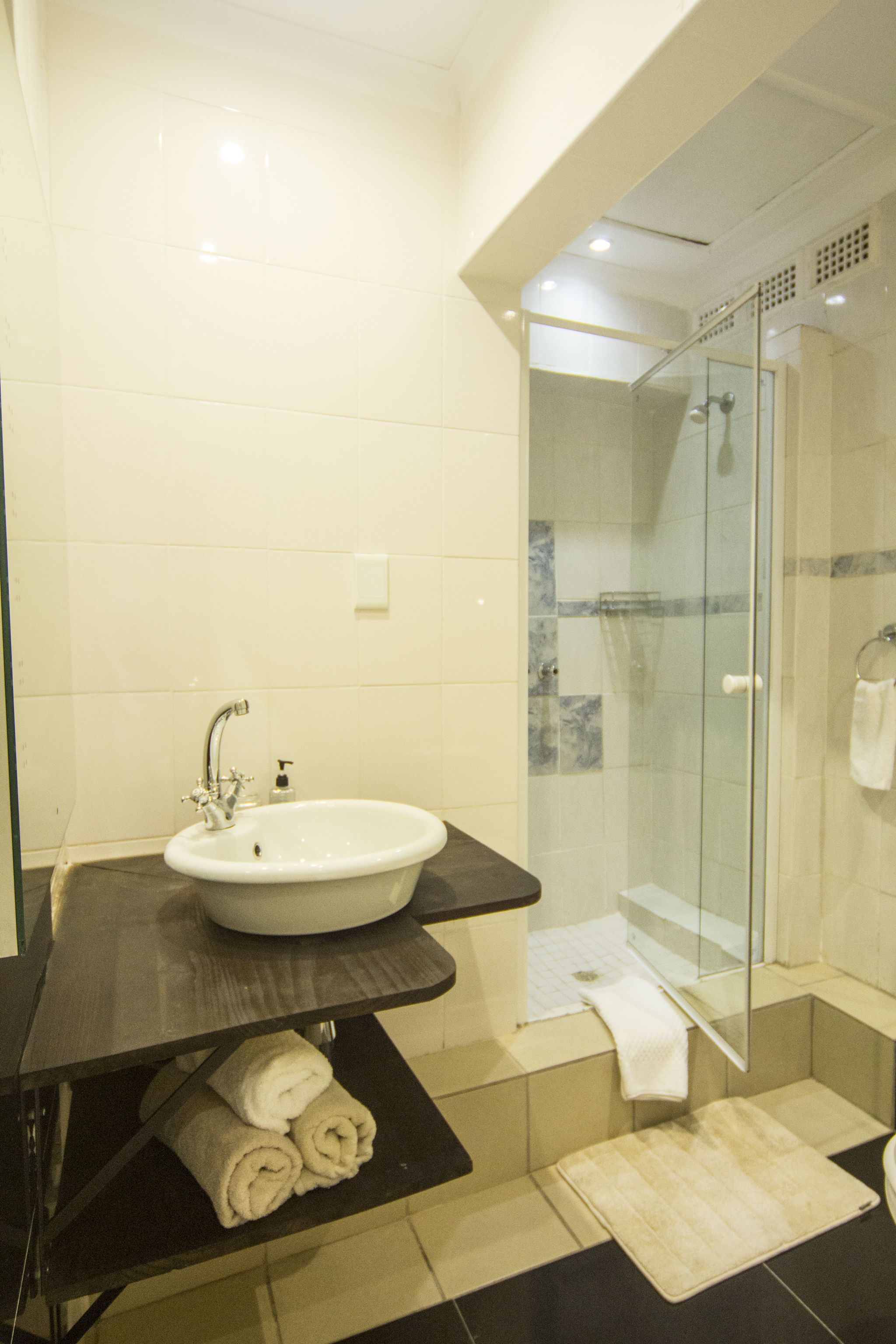 A modern bathroom with a glass-enclosed shower, a sink with a dark countertop, and neatly rolled towels on a shelf.
