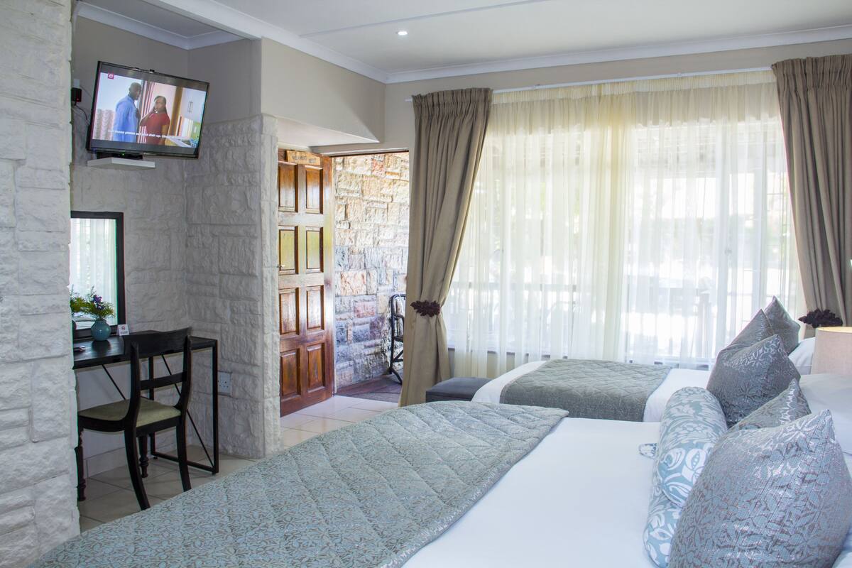 A hotel room with a stone wall, a television, a desk with a chair, two beds with patterned bedding, and a large window with curtains.