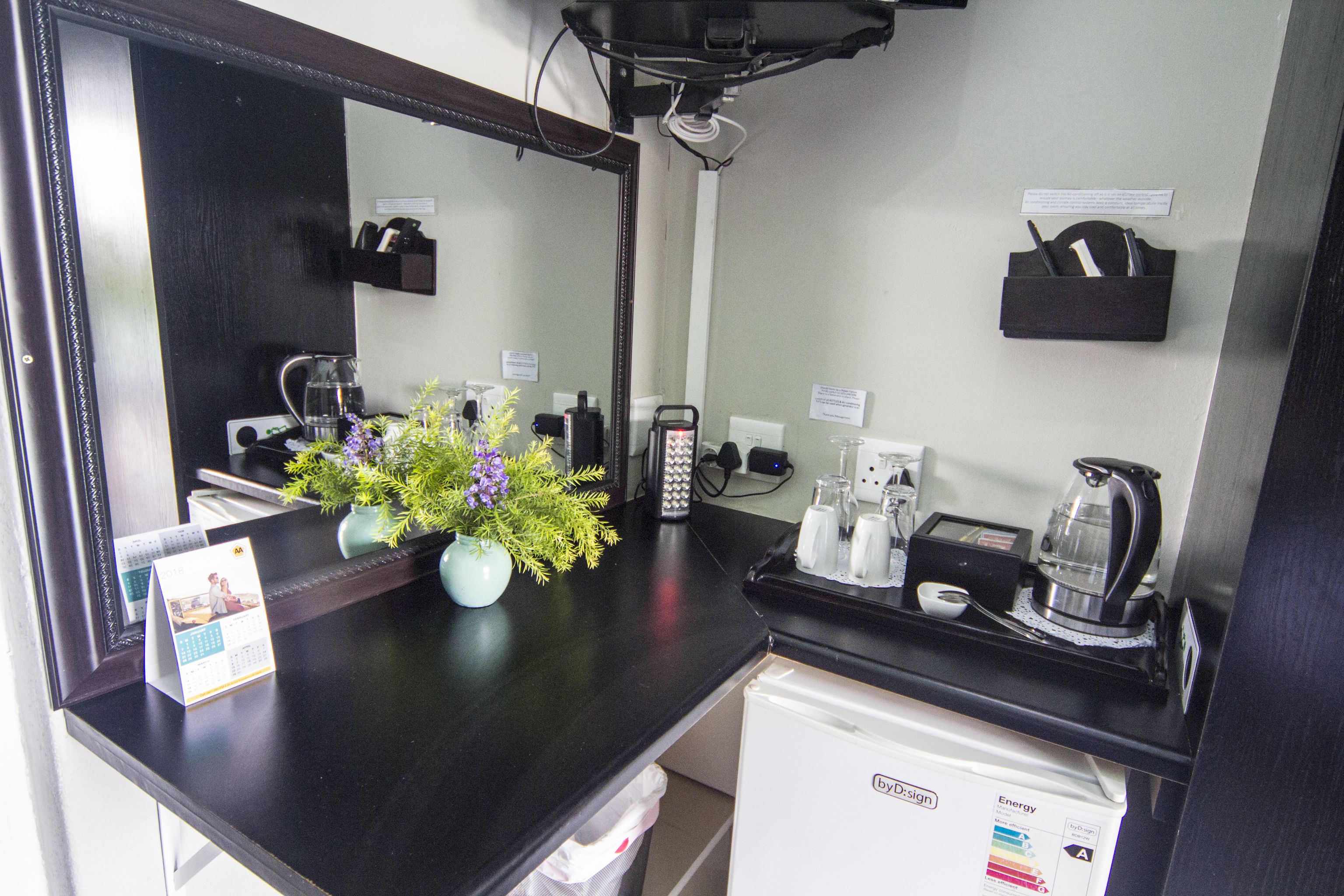 Habitación de hotel con una encimera negra, un refrigerador blanco, un espejo, un jarrón con flores, un calendario y una cafetera.