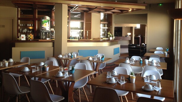 Un restaurant avec des tables en bois et des chaises blanches, un coin bar et une vue sur la cuisine.