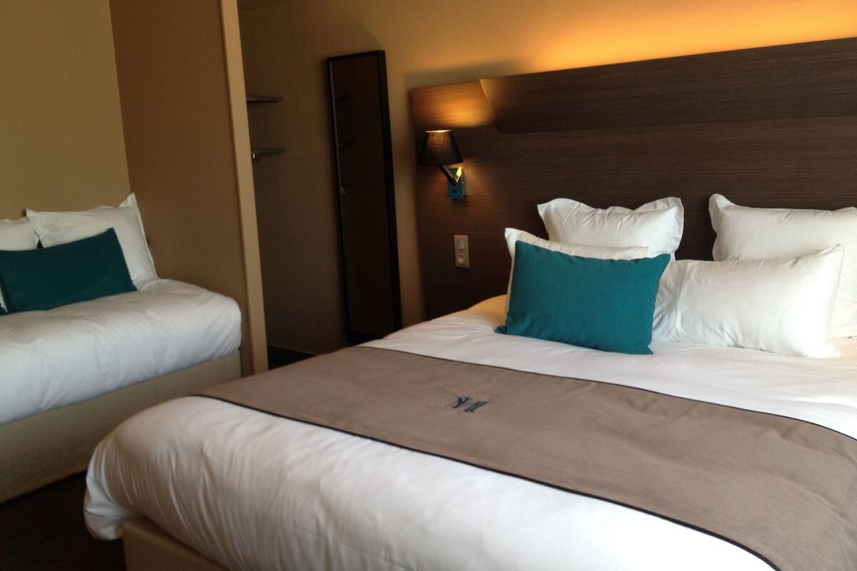 A hotel room with two beds, each with white linens and teal pillows. There is a wooden headboard and a wall-mounted light fixture.
