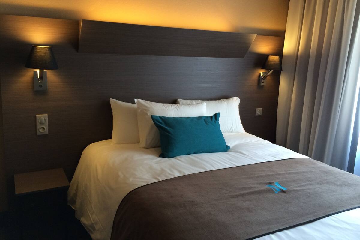 A hotel room with a bed, two wall-mounted lamps, and a window with curtains.