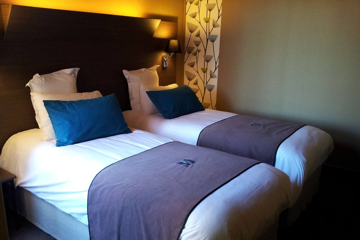 A hotel room with two beds, each with white linens and blue pillows. There is a dark headboard, a wall-mounted lamp, and a decorative wall panel.