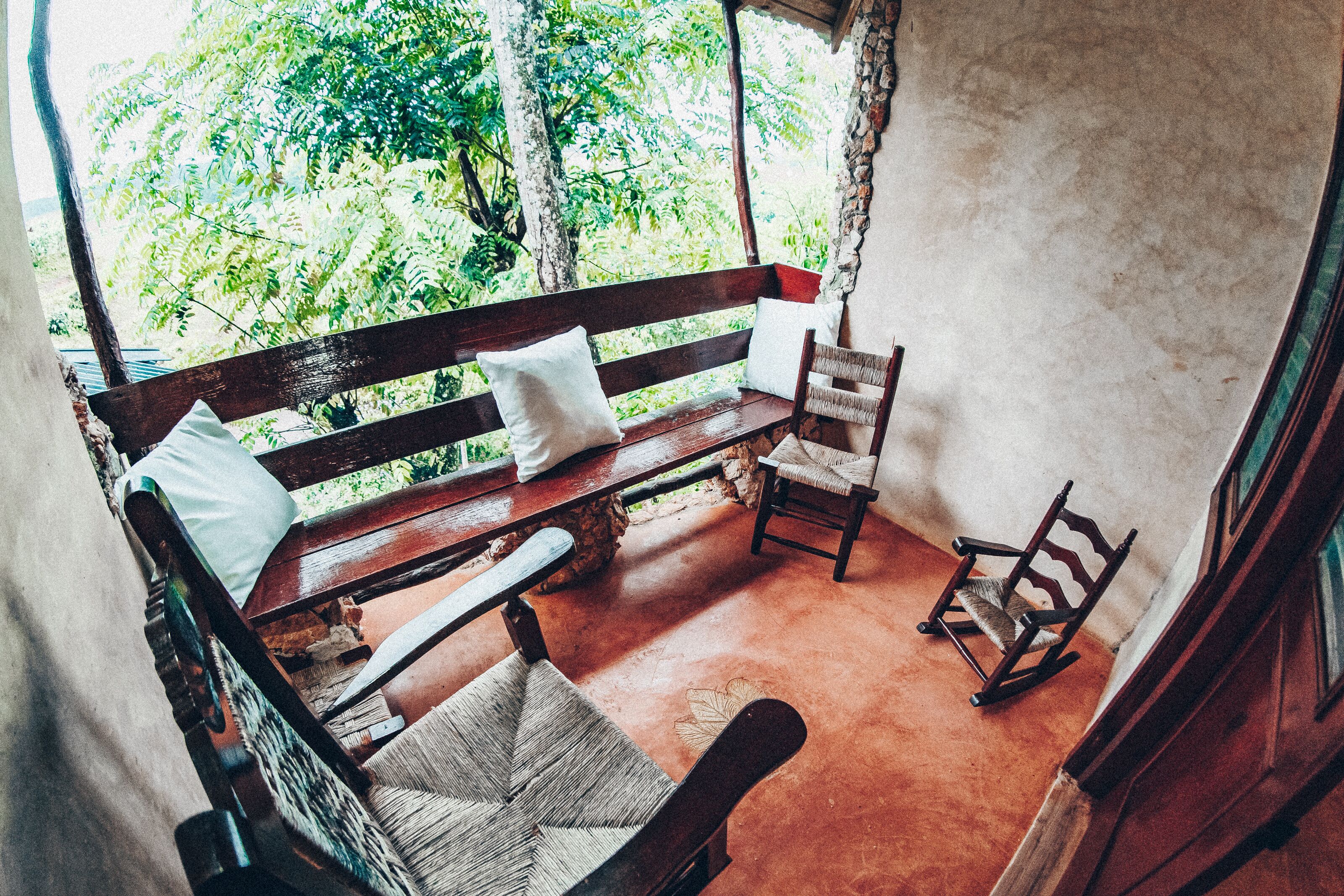A wooden bench with a cushion, a wicker chair, and a rocking chair in a rustic room with a view of greenery outside.