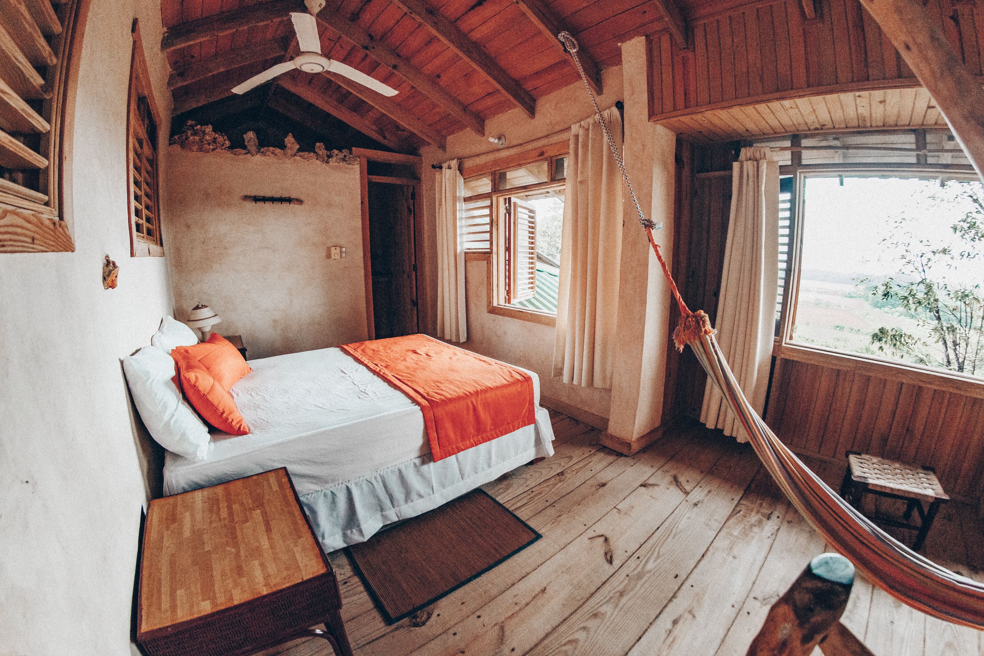 A bedroom with a bed, wooden floor, a hammock, a small table, and a window with a view.