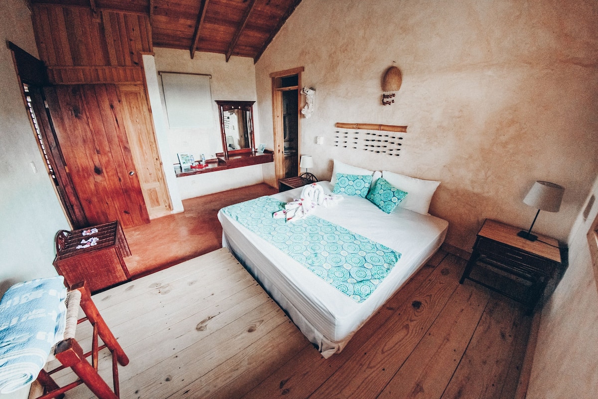 A bedroom with a wooden floor, a bed with patterned bedding, a nightstand, a bedside table, a wooden wardrobe, and a mirror.