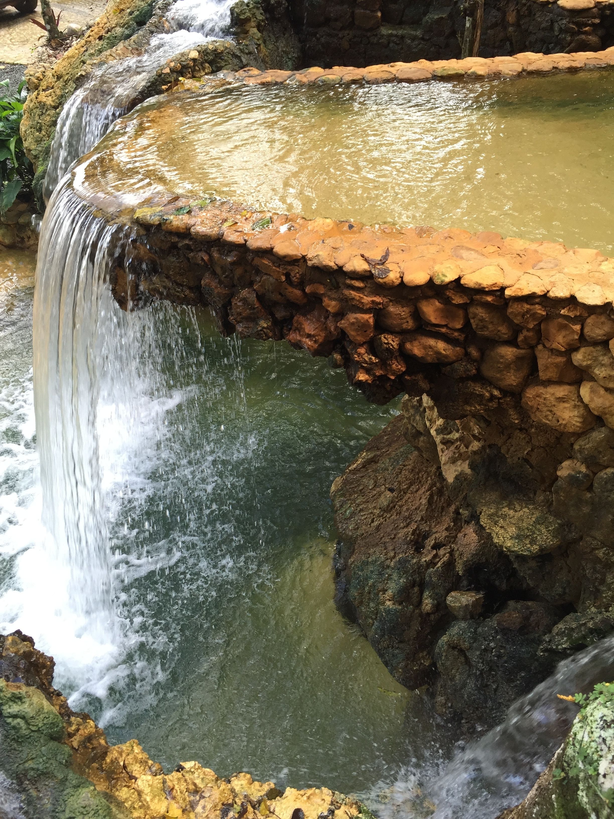 Una fuente de piedra con una cascada que desemboca en un estanque, rodeada de rocas y vegetación.