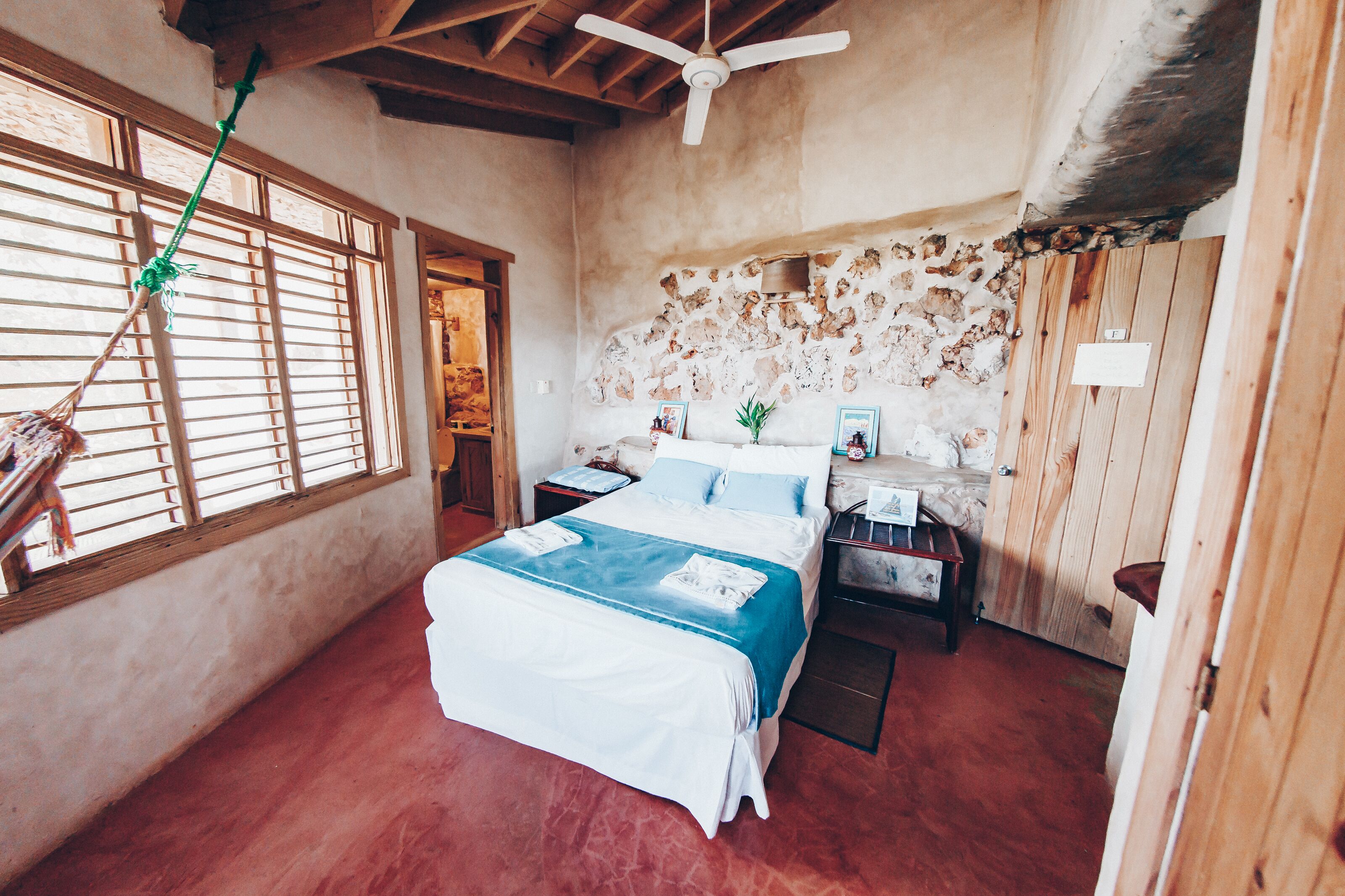 Ein Schlafzimmer mit einem Bett, einem Holzkopfende, einem Deckenventilator, einem Fenster mit Jalousien und einer Steinwand.