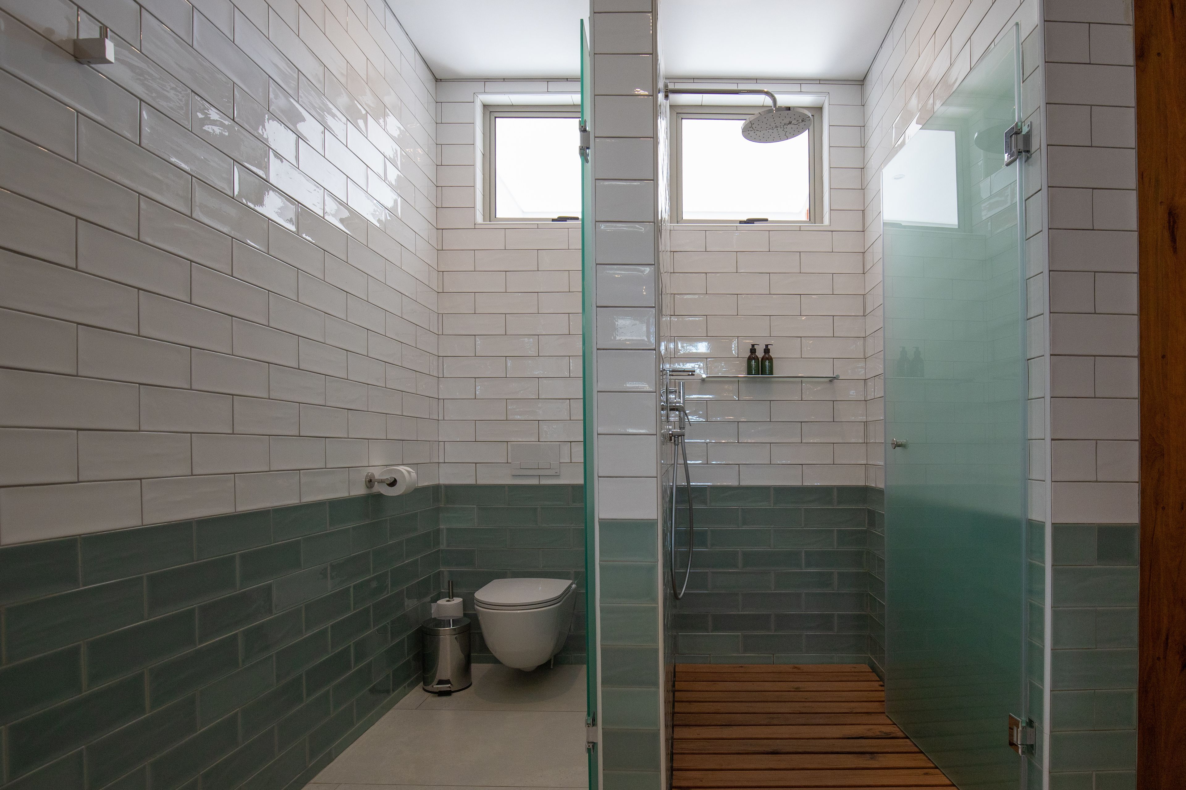 A bathroom with white and green tiles, a glass shower enclosure, a wooden bath mat, and a toilet.