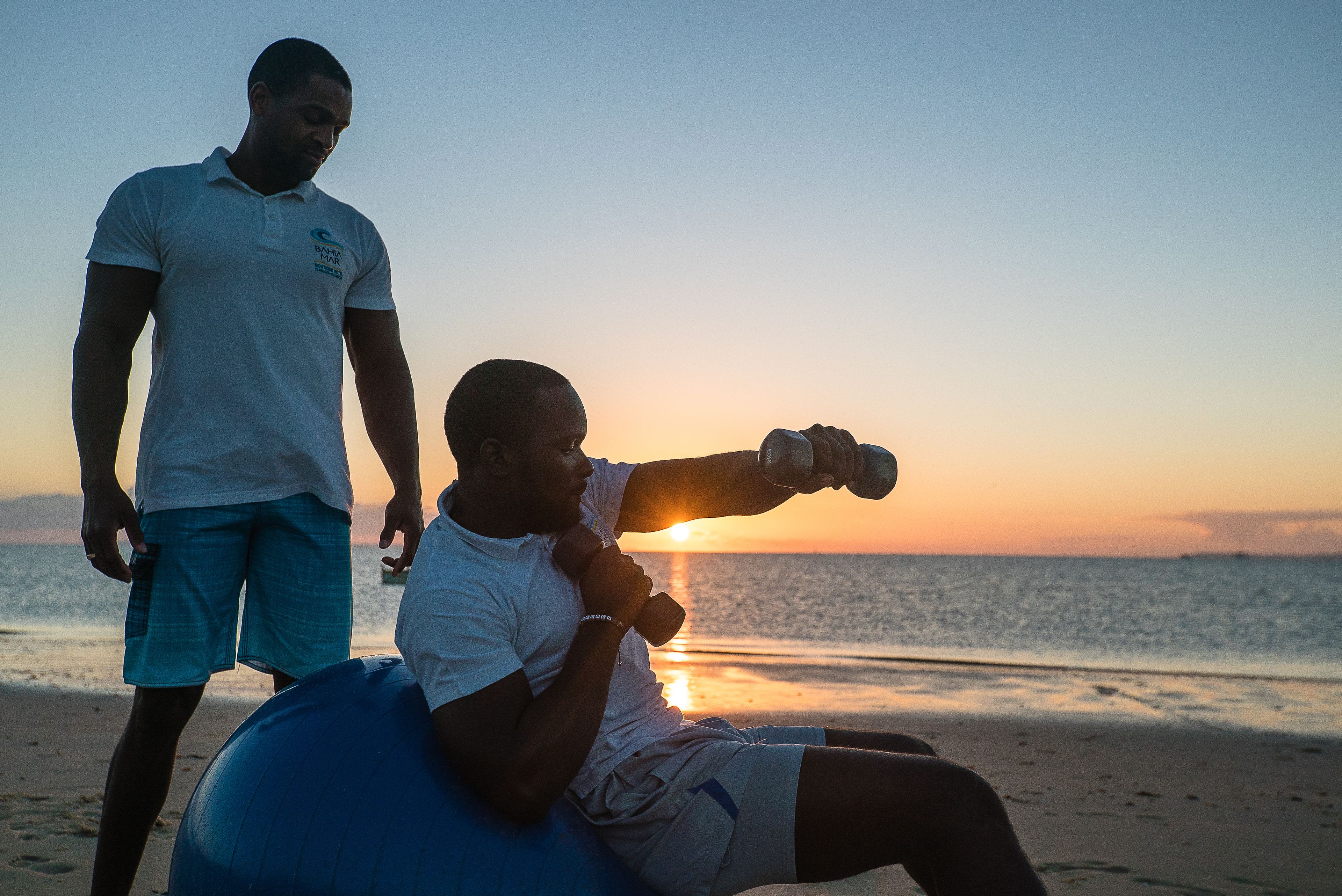 Una persona sentada en una pelota azul de ejercicios en la playa al atardecer. Otra persona está cerca.
