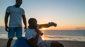 Una persona sentada en una pelota azul de ejercicios en la playa al atardecer. Otra persona está cerca.