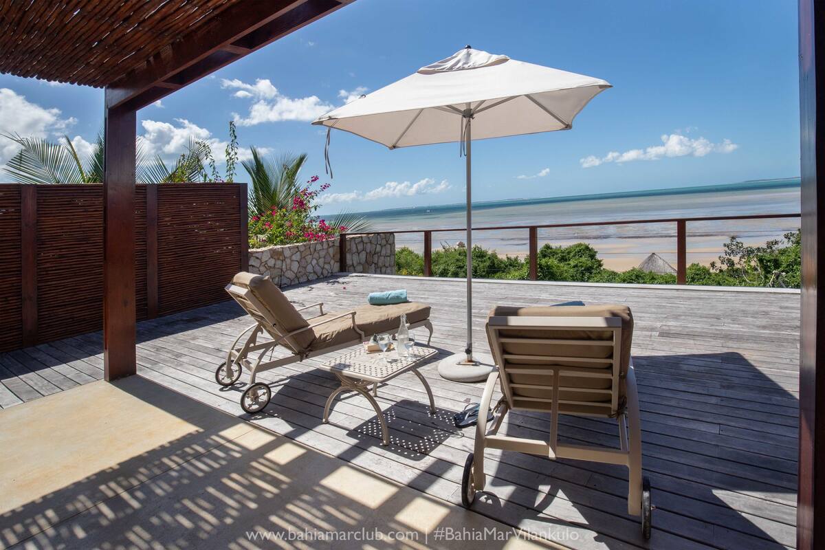 Una terraza de madera con dos sillones y una mesa, con vista a la playa.