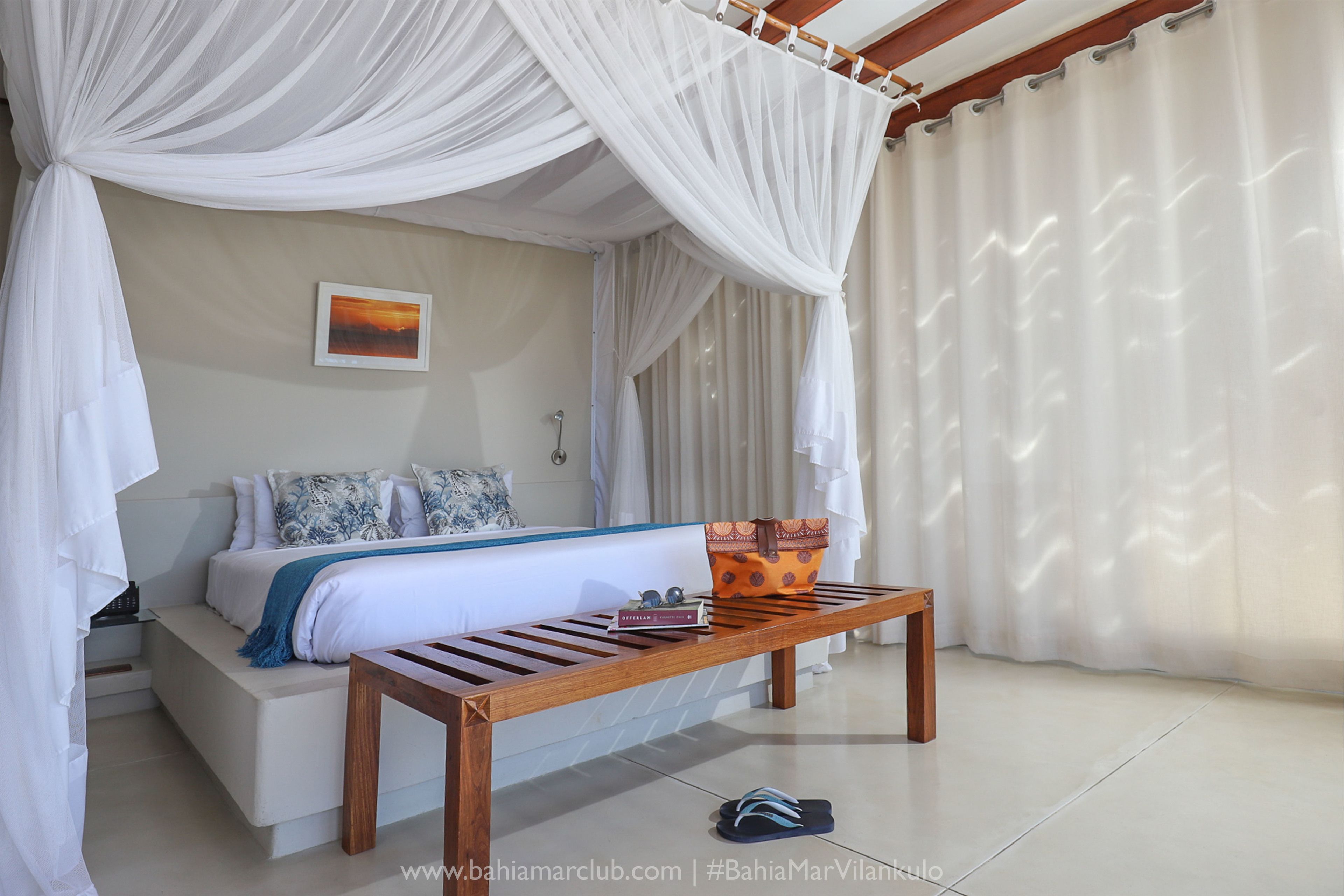 A bedroom with a bed, a bench, a pillow, a blanket, a pillowcase, a wall-mounted light, and a framed picture.