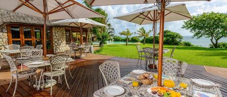An outdoor dining area with tables and chairs, umbrellas, and a view of the sea.