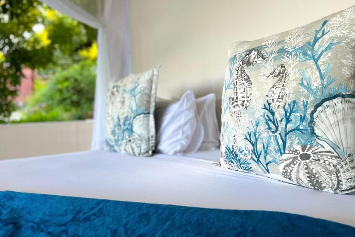 Een bed met een wit-blauwe dekbedovertrek, twee kussens met een blauw-wit patroon, en uitzicht op groen buiten.