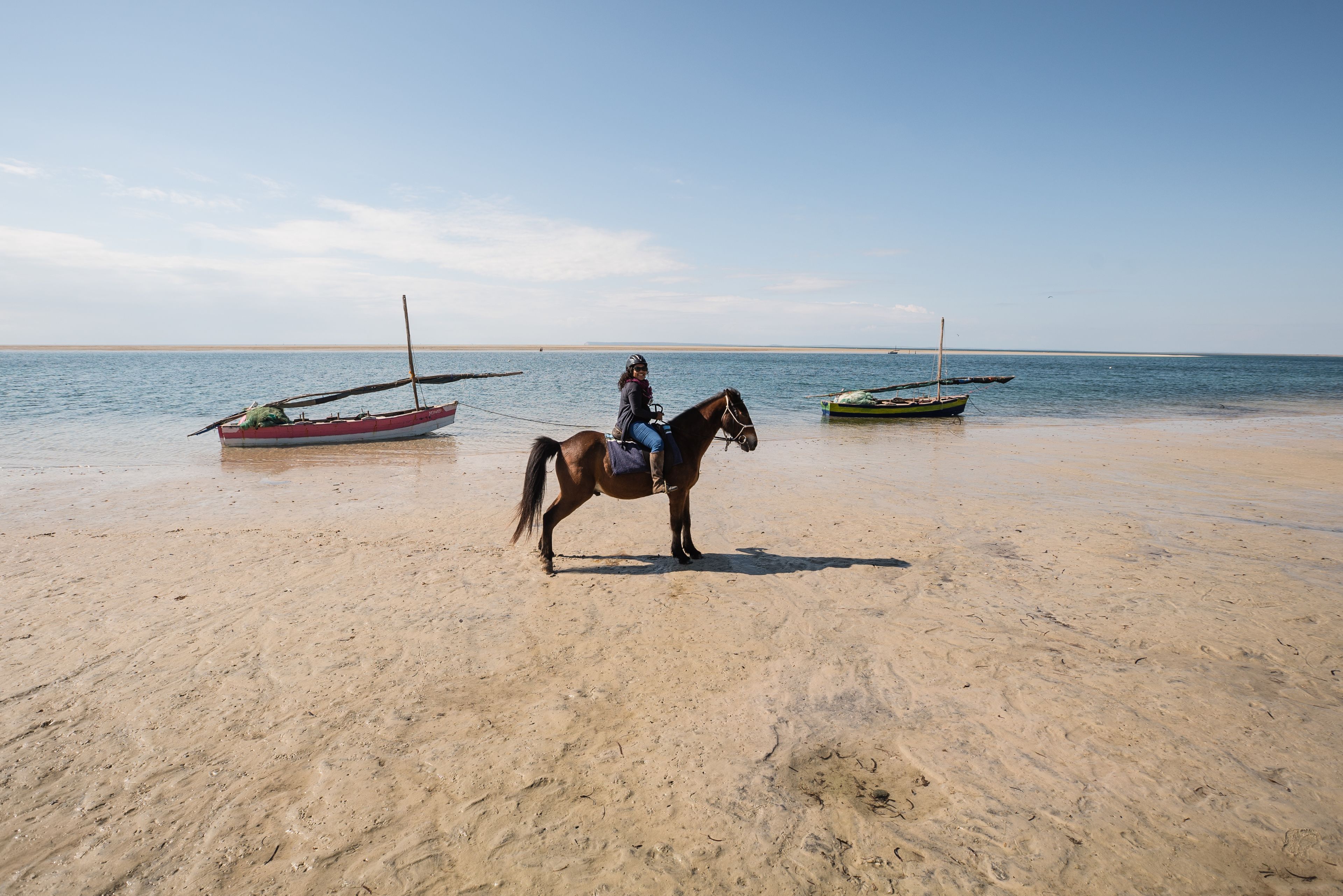 A sandy beach with a horse and rider, and two boats in the background.
