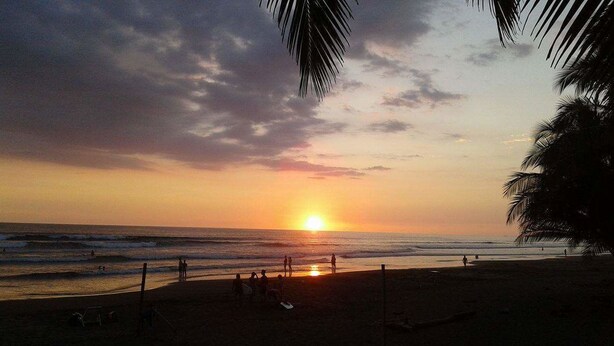 Una playa al atardecer, con el sol poniéndose sobre el océano, palmeras y gente en la arena.