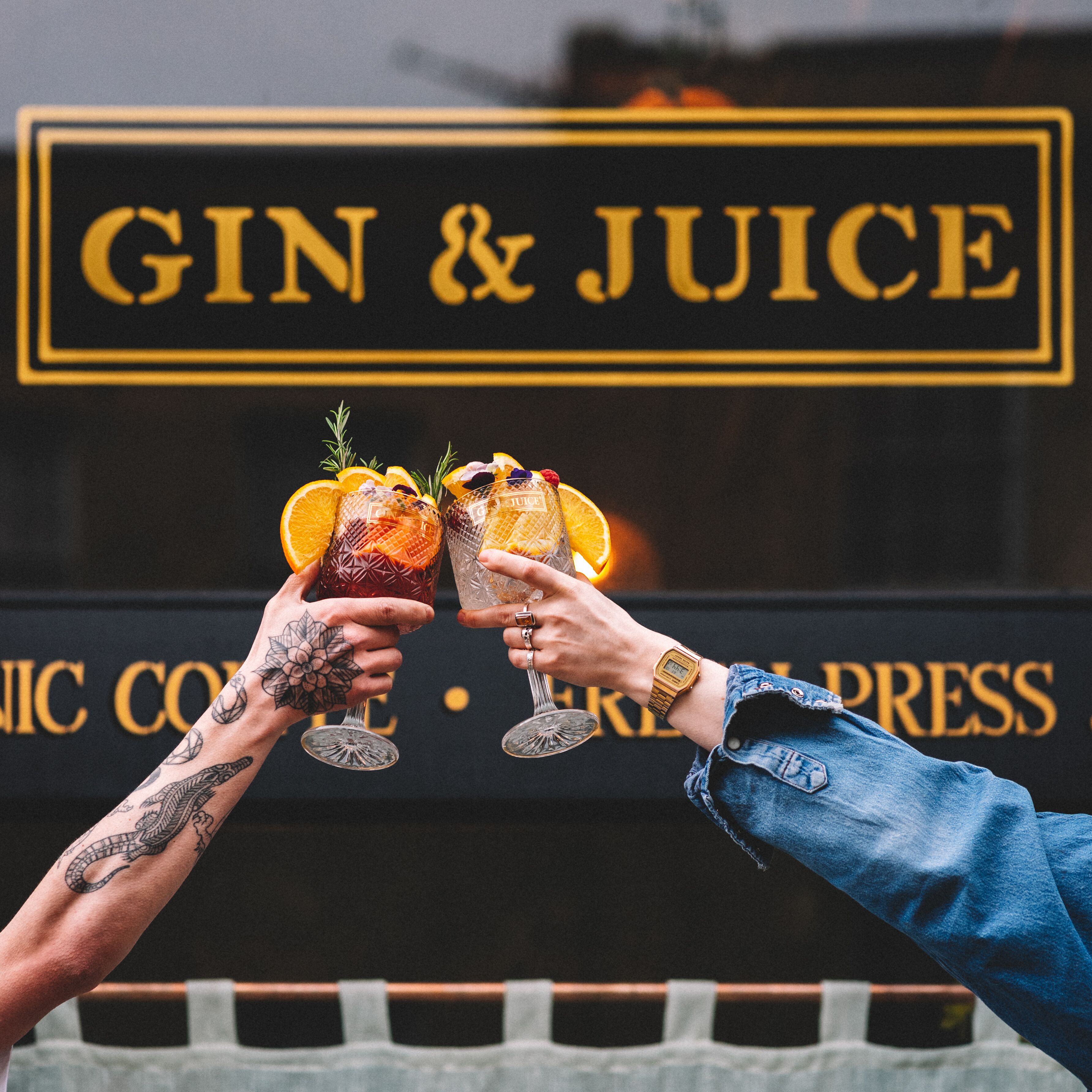 Two people toasting with drinks in front of a sign that reads "GIN & JUICE".