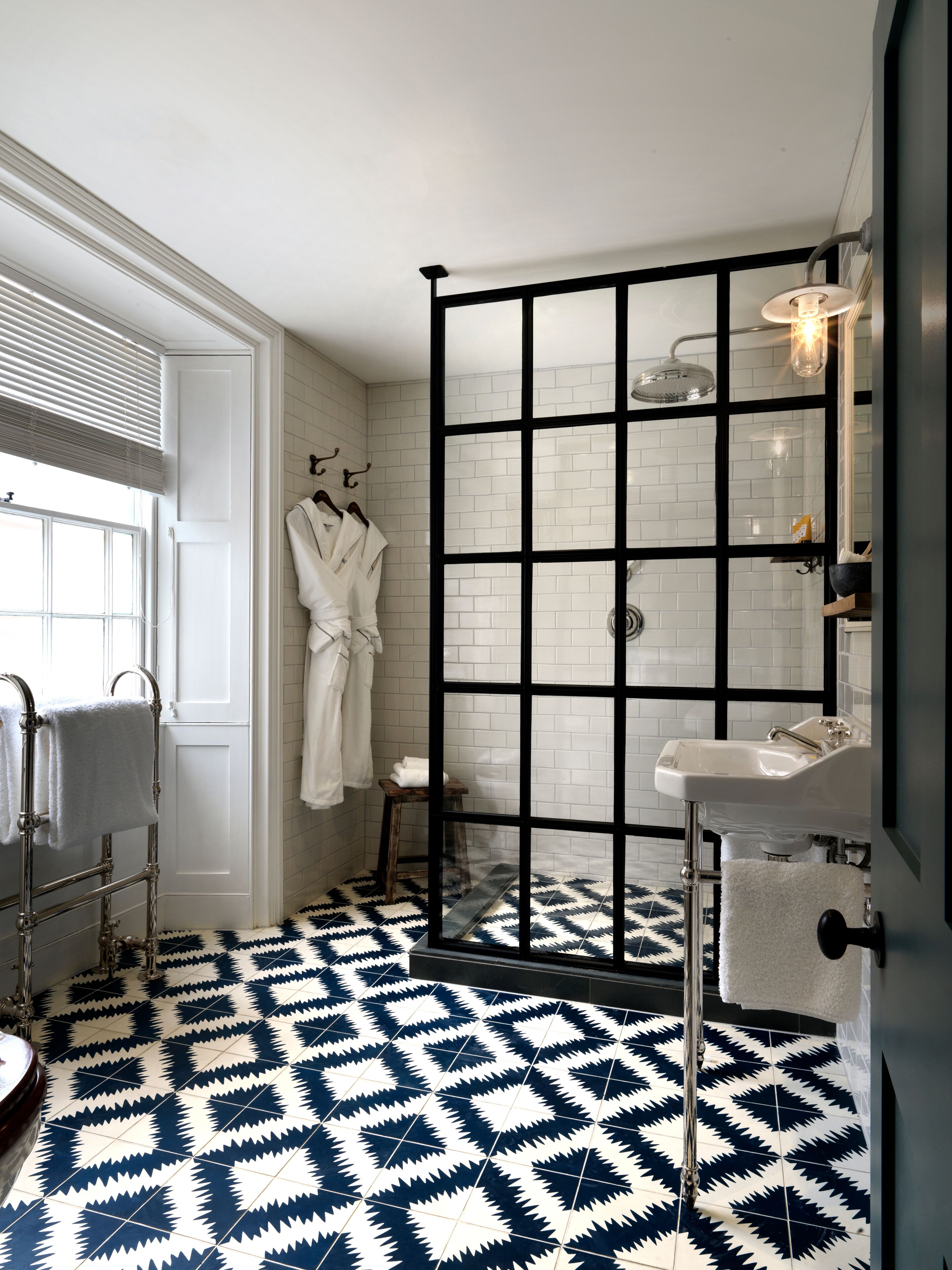 Un baño con mampara de ducha de vidrio con marco negro, paredes con azulejos blancos, un lavabo blanco y piso estampado.