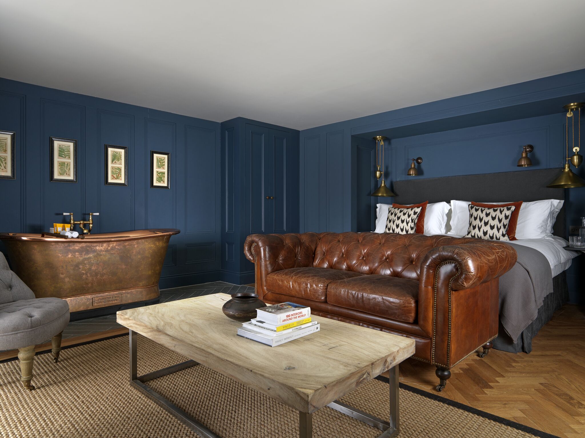 A room with a brown leather sofa, a wooden coffee table with books, a vintage bathtub, and a bed with patterned pillows.