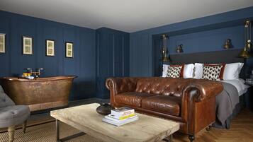 A room with a brown leather sofa, a wooden coffee table with books, a vintage bathtub, and a bed with patterned pillows.