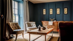 Een kamer met een groot raam, een open haard, een tafel met waterkoker en kunstwerken in lijsten aan de muur.