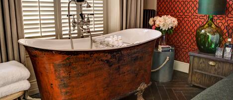 A vintage clawfoot bathtub in a bathroom with a patterned wallpaper, a green vase, and a small wooden side table.