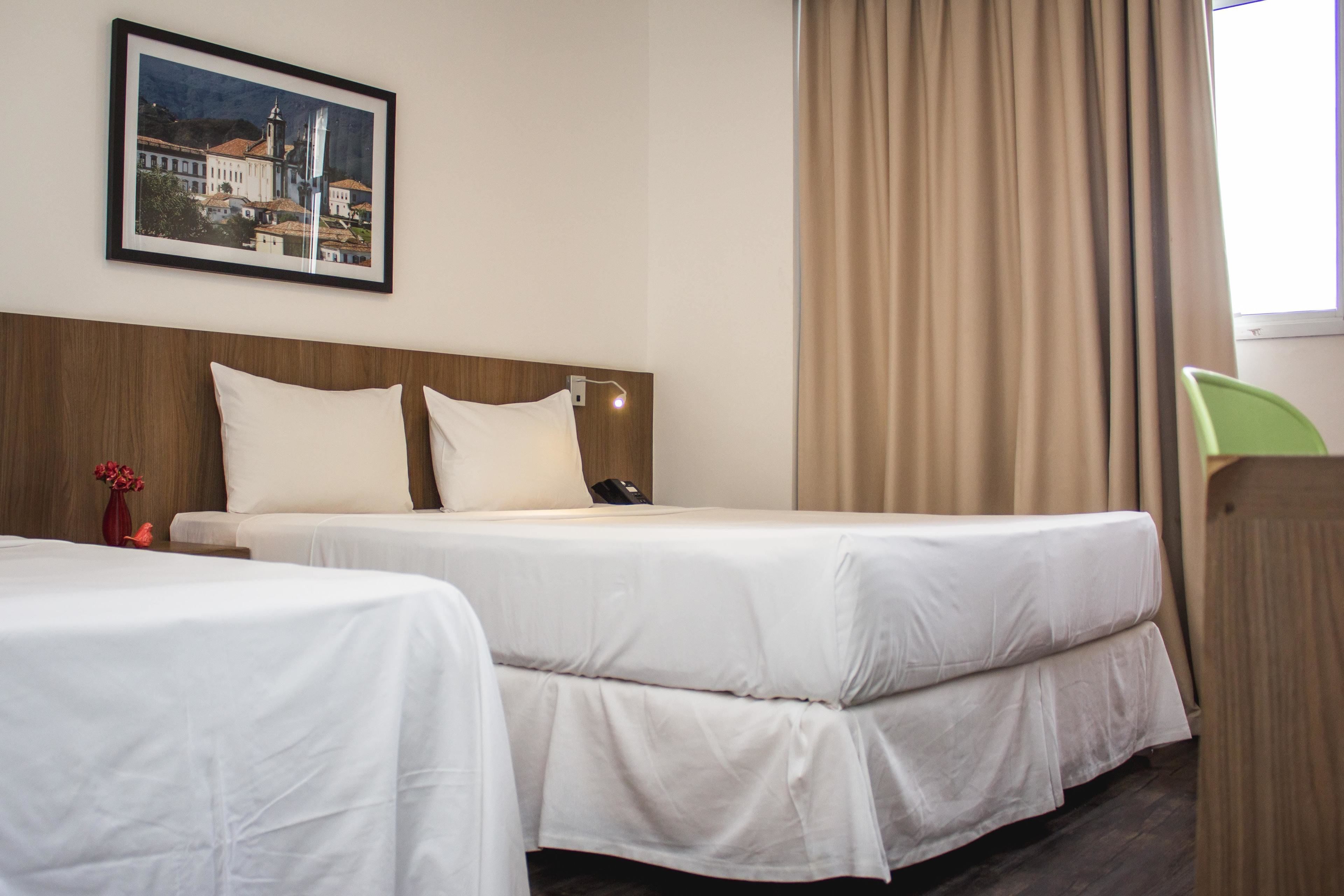Quarto de hotel com duas camas, uma cabeceira de madeira, um quadro na parede e uma janela com cortinas.