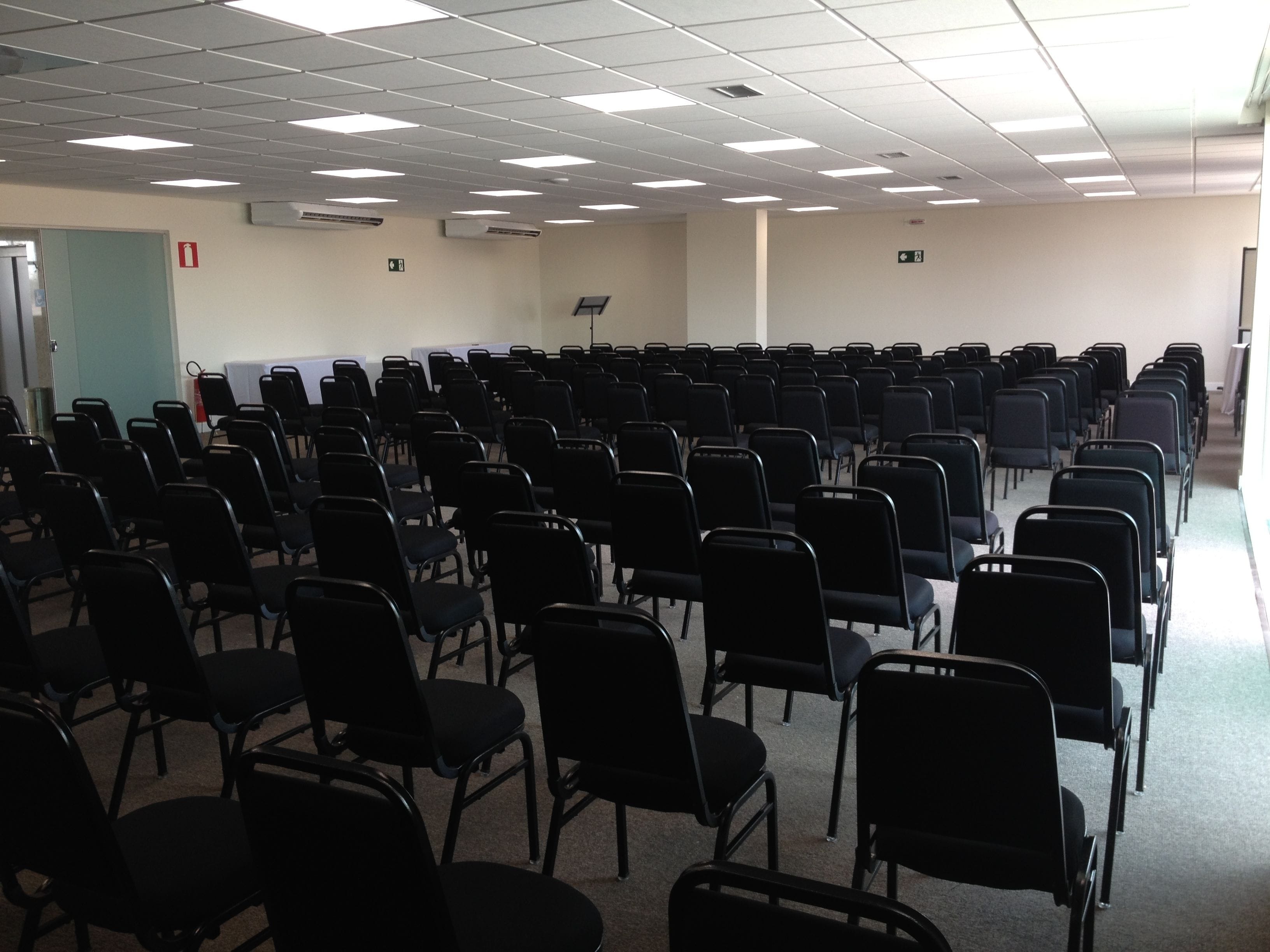 Un salón de conferencias con filas de sillas negras, un techo blanco con luces fluorescentes y una pizarra en la pared.