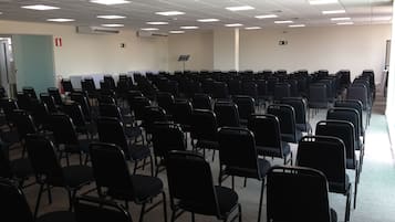 Un salón de conferencias con filas de sillas negras, un techo blanco con luces fluorescentes y una pizarra en la pared.