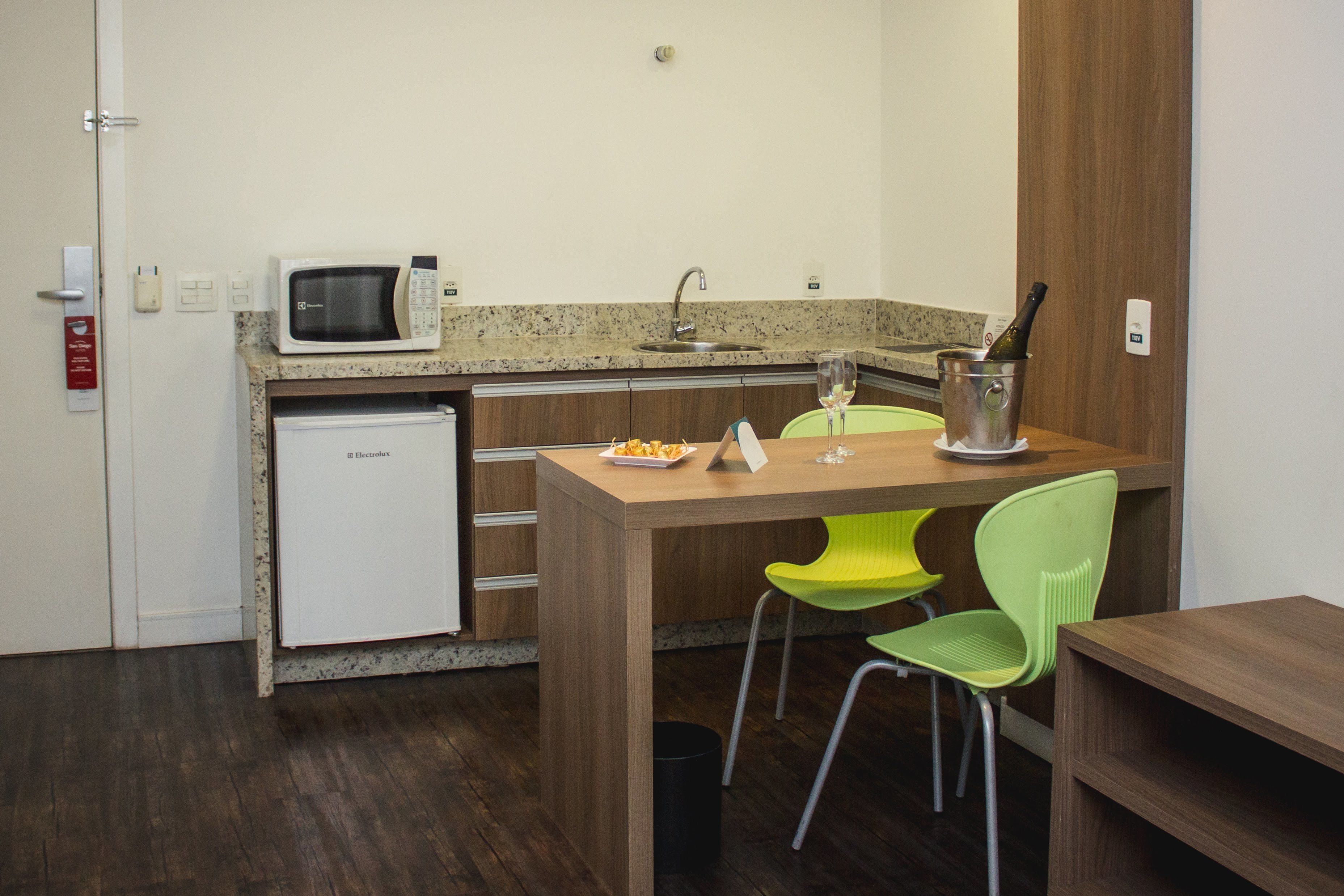 Una cocina compacta con microondas, lavamanos y un refrigerador pequeño. Hay una mesa de comedor con dos sillas y una botella de champán.