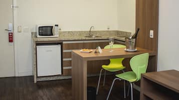Una cocina compacta con microondas, lavamanos y un refrigerador pequeño. Hay una mesa de comedor con dos sillas y una botella de champán.
