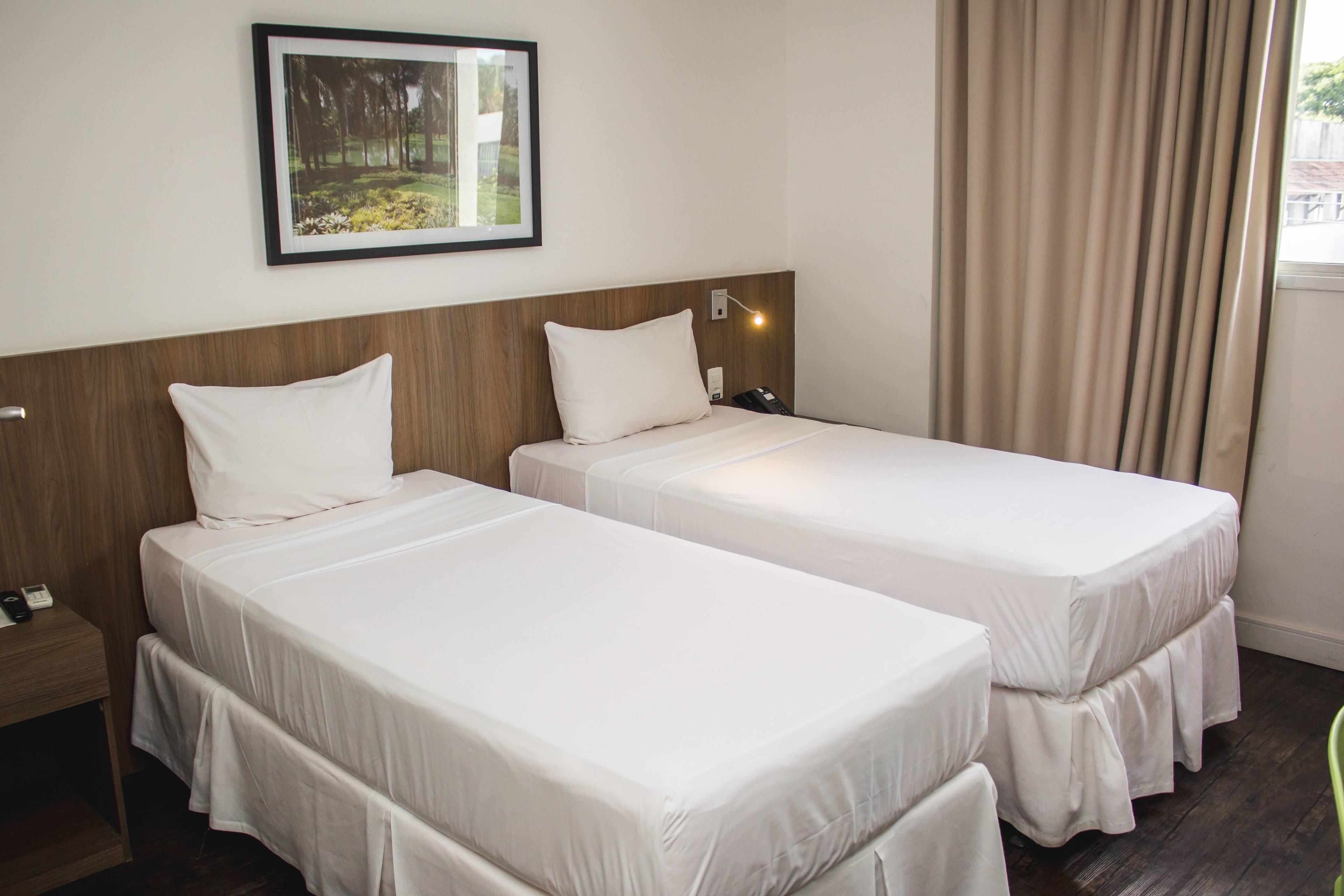 Quarto de hotel com duas camas de solteiro, cabeceira de madeira, mesa de cabeceira com telefone, quadro na parede e janela com cortinas.
