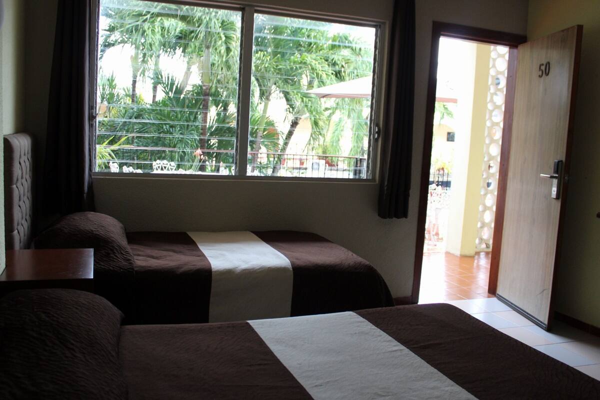 A room with two beds, a window with blinds, a door, and a view of greenery outside.