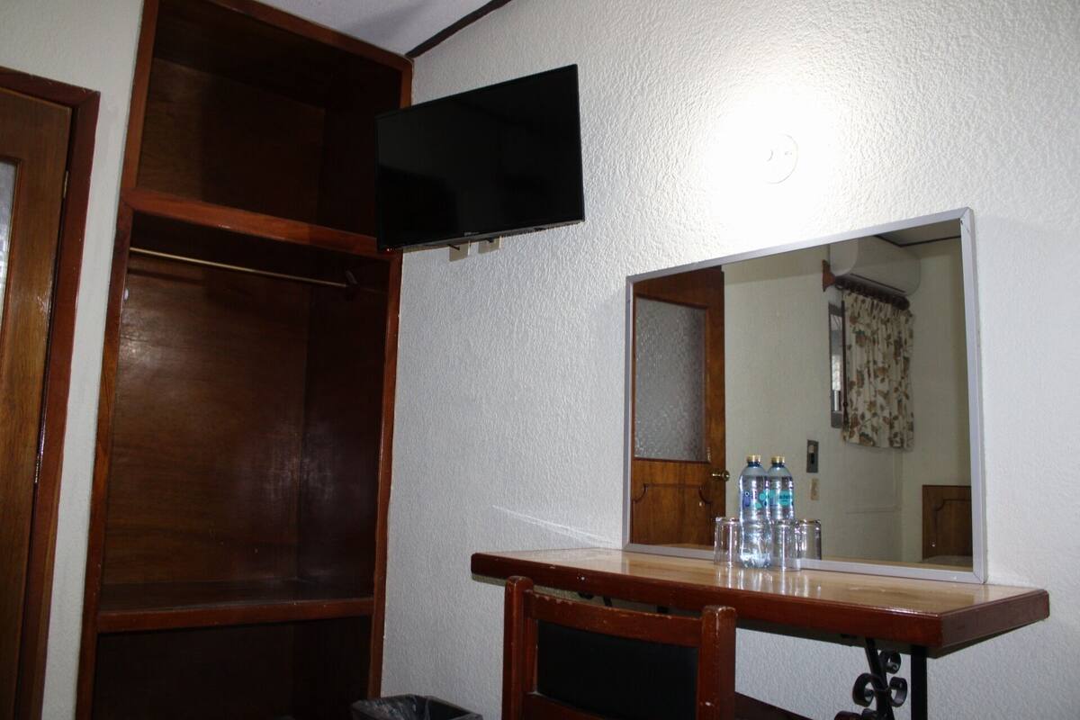 A room with a wooden wardrobe, a flat-screen TV mounted on the wall, a mirror above a wooden table with water bottles, and a door.