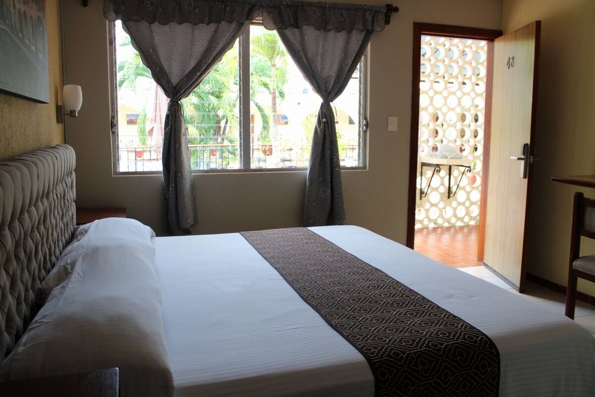 A hotel room with a bed, a window with curtains, a door leading to a balcony, and a wall-mounted light.