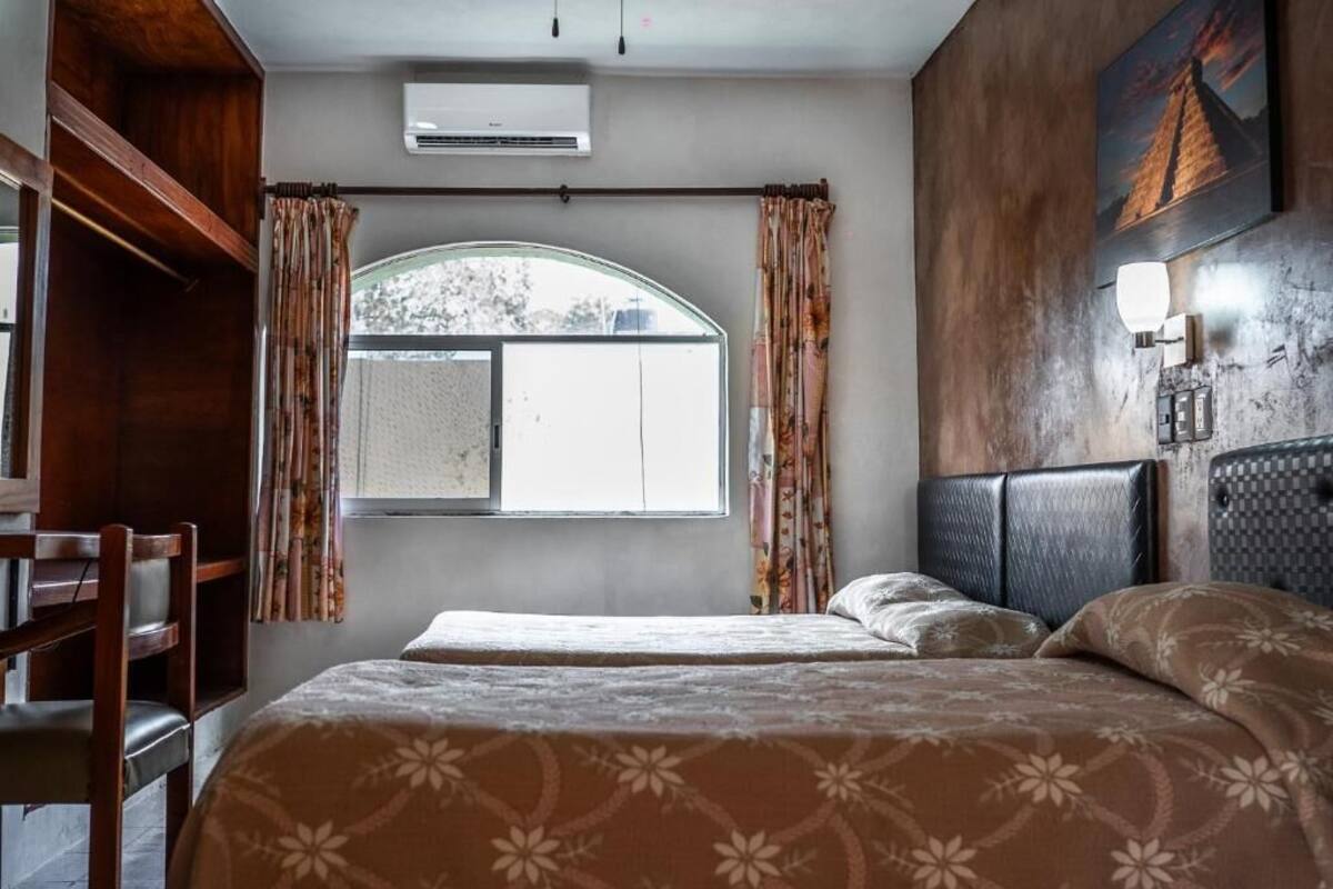 A bedroom with a large bed, a wooden wardrobe, a window with curtains, and a wall-mounted air conditioner.