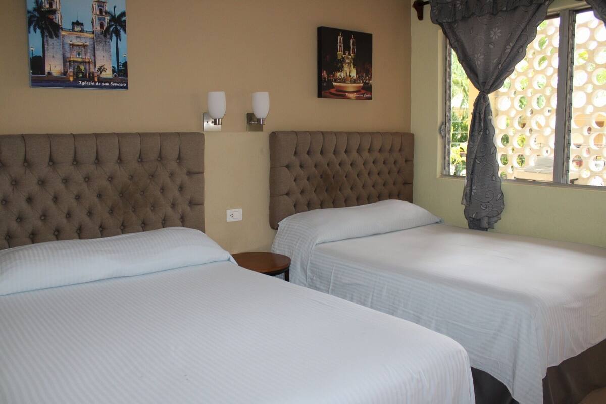 A hotel room with two beds, a large tufted headboard, a poster of a building, and a window with a patterned curtain.