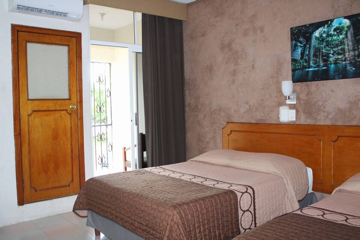 A bedroom with a wooden door, a bed with a patterned bedspread, a wall-mounted light, and a painting of a waterfall.