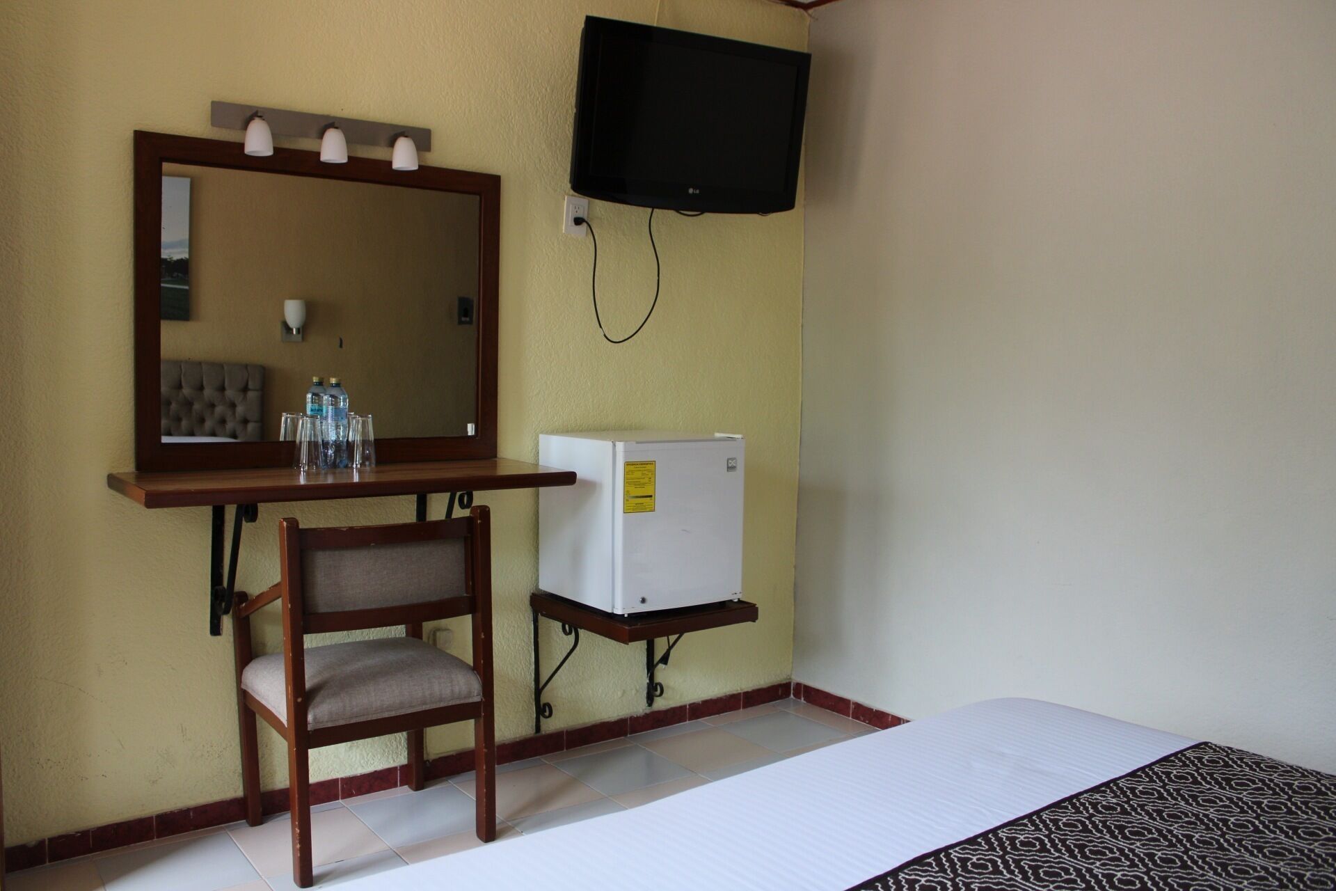 Habitación de hotel con una cama, una silla de madera, una mesita con vasos y botellas, un espejo, un televisor y un refrigerador.