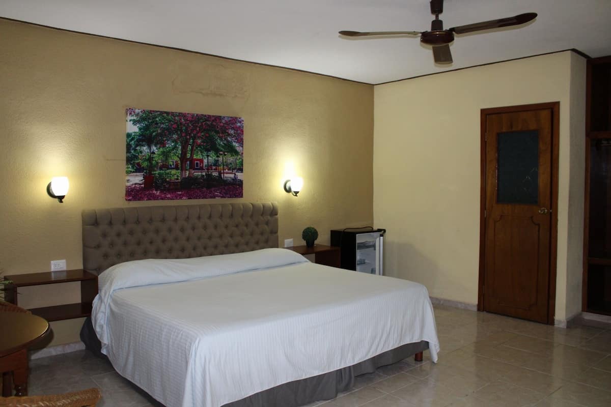 A hotel room with a bed, two wall-mounted lights, a ceiling fan, a door, and a painting on the wall.