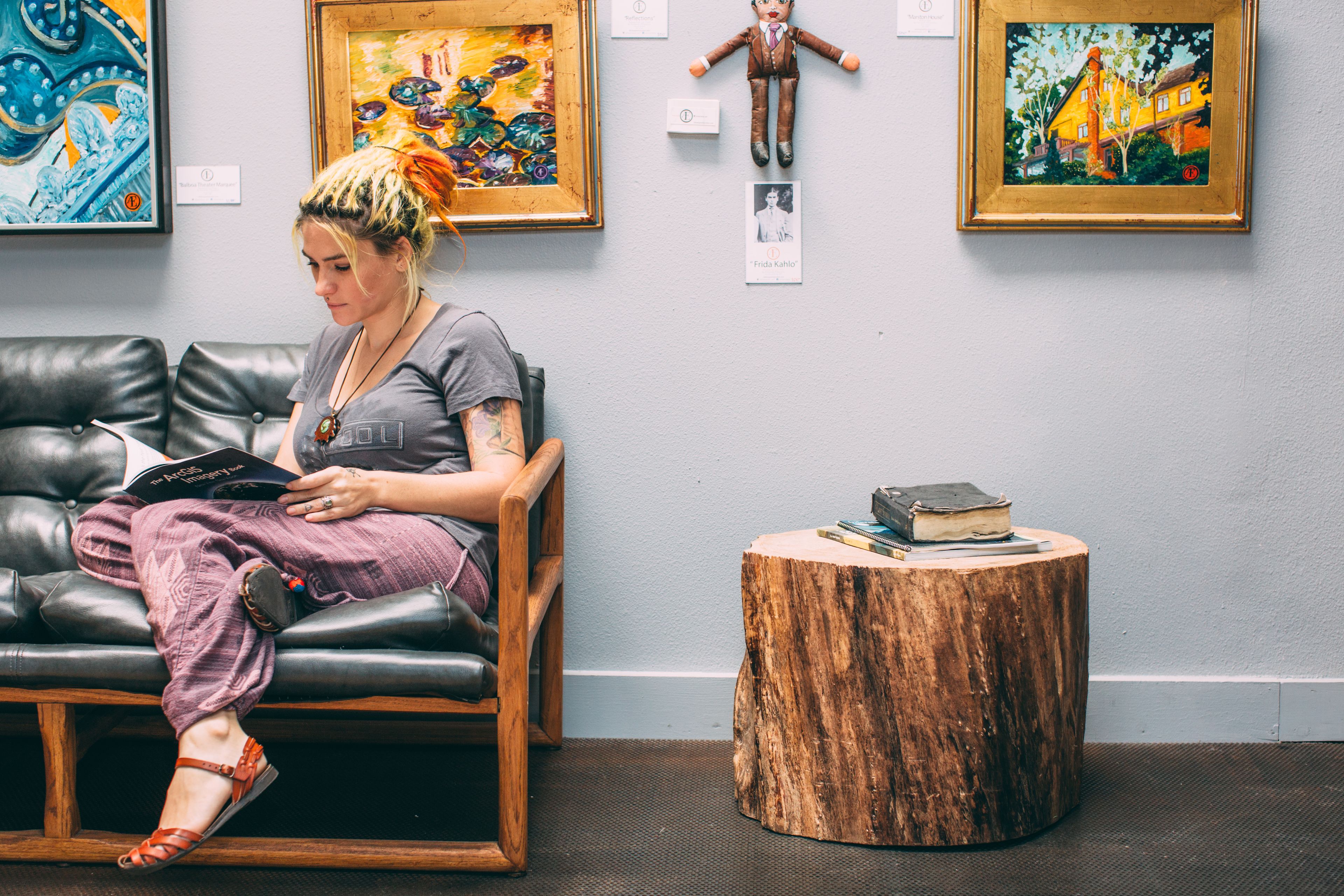 A person sitting on a leather couch in a room with a wooden side table, a framed painting on the wall, and a small wooden stool.