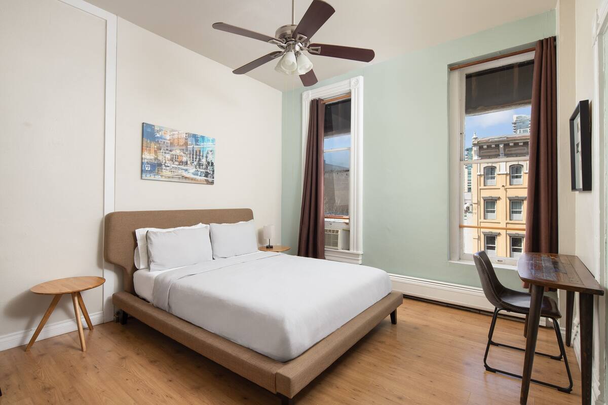 Un dormitorio con cama, ventilador de techo, ventana con cortinas, una mesita y una silla.