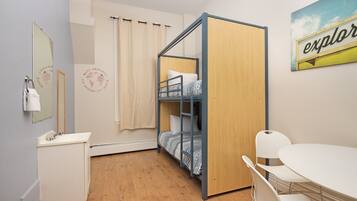 A dormitory room with a bunk bed, a small desk with a mirror, a white table, and a painting on the wall.
