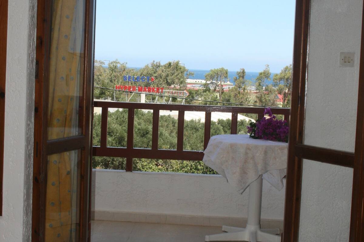 A balcony with a view of the sea, a table with a tablecloth, and a rolled-up towel on the floor.