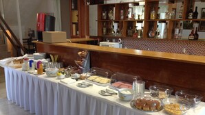 Un buffet proposant divers plats et accompagnements, disposés sur une table, avec une machine à café et un bar où l’on aperçoit des bouteilles à l’arrière-plan.