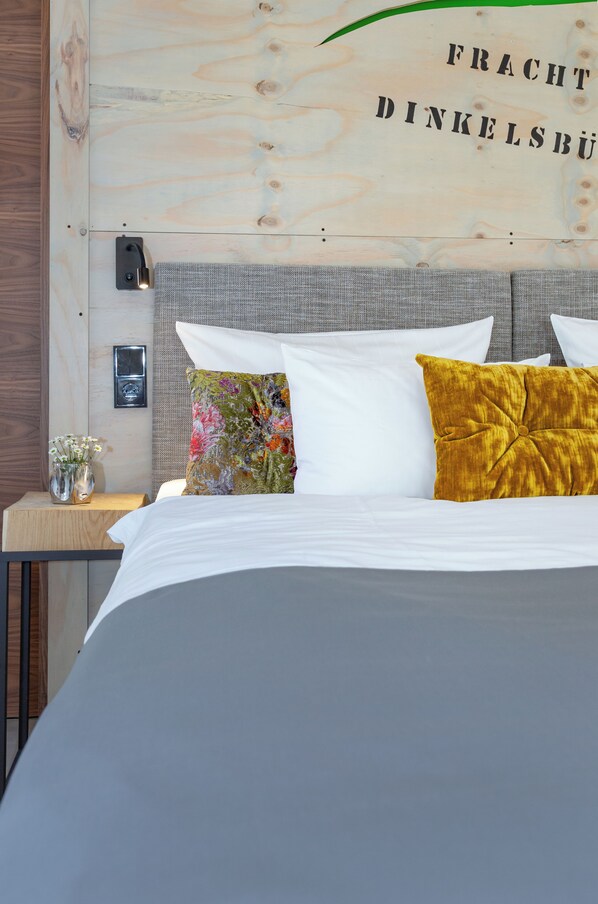 Una cama bien hecha con almohadas blancas y mostaza, un jarrón pequeño con flores y una pared con panel de madera y texto.