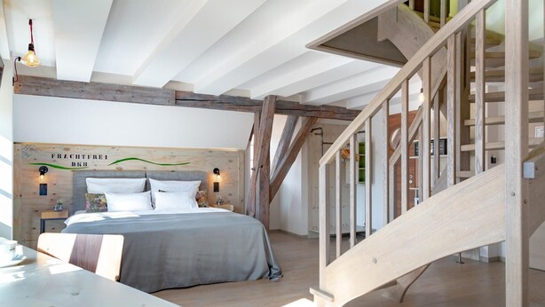 A bedroom with a wooden headboard, a bed with white bedding, a wooden staircase, and a view of the outside. - Hotel Luise & Luisenhof (Dinkelsbuehl)