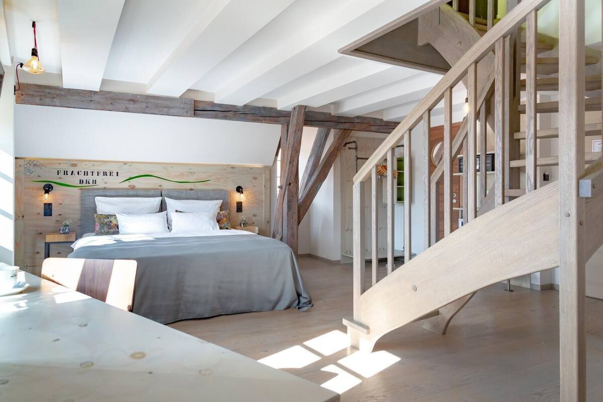 Un dormitorio con cabecera de madera, cama con ropa de cama blanca, una escalera de madera y vistas al exterior.