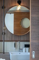 Un baño moderno con un tocador de madera, un espejo redondo, un lavabo blanco y una lámpara sobre el espejo.